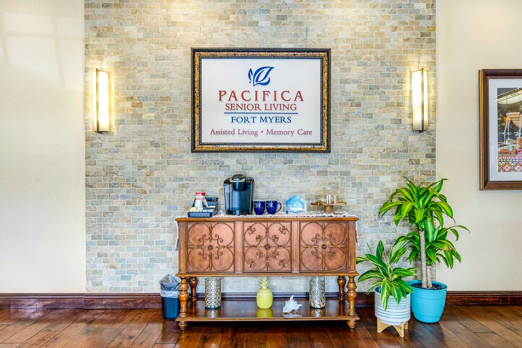 A welcoming coffee station with a wooden cabinet in an elegant interior setting featuring the sign 'Pacifica Senior Living' on the wall.