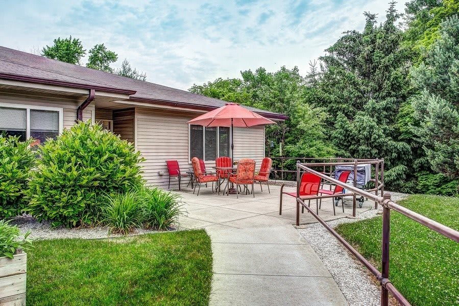 A cozy backyard patio with red chairs and an umbrella next to a house, surrounded by lush greenery.