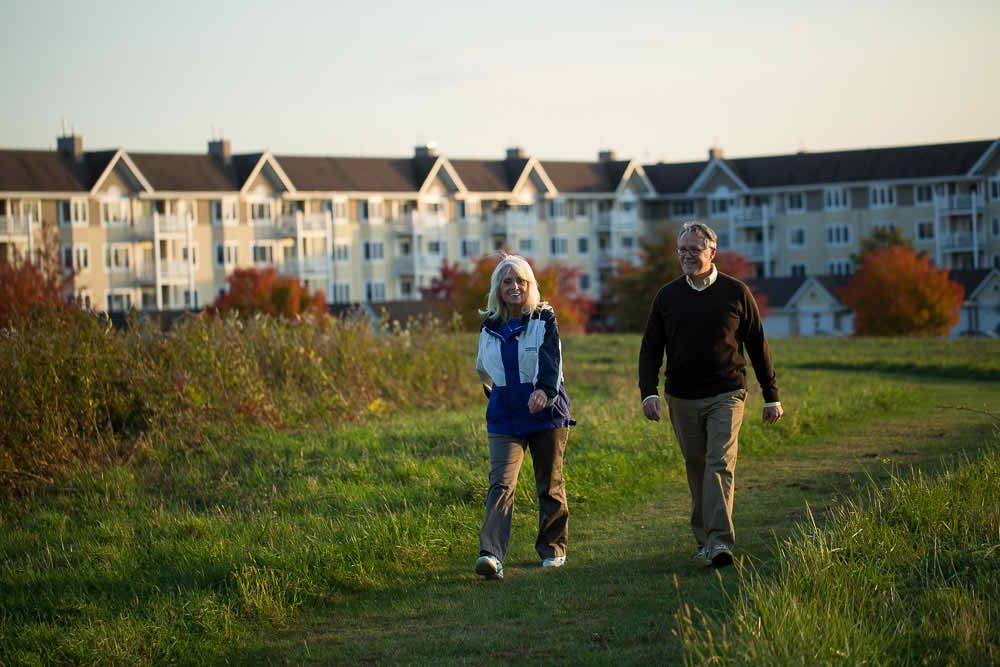 A couple walking outdoors on a grassy path with buildings in the background.