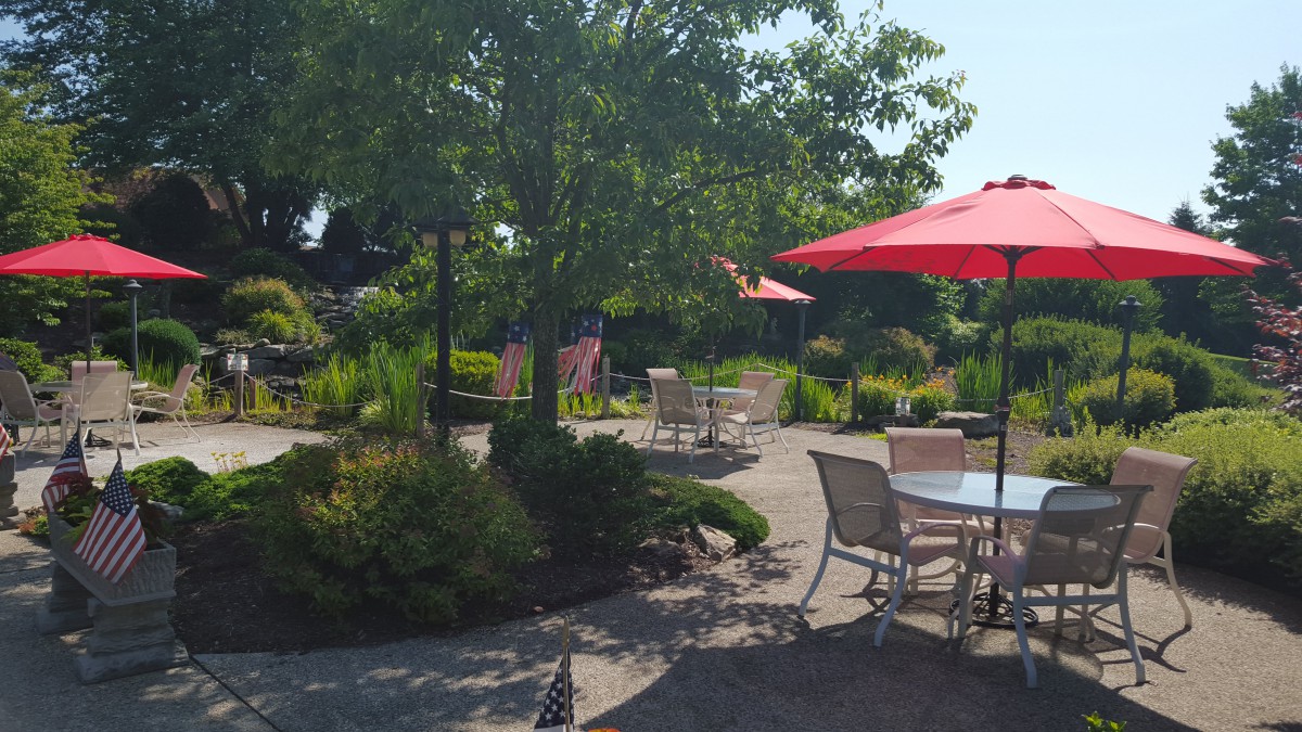 A sunny garden patio with chairs, tables, and red umbrellas surrounded by lush greenery.