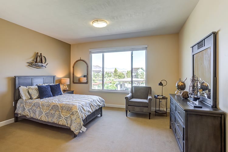 A cozy bedroom with a large window, featuring a bed, armchair, dresser, and decorative elements.