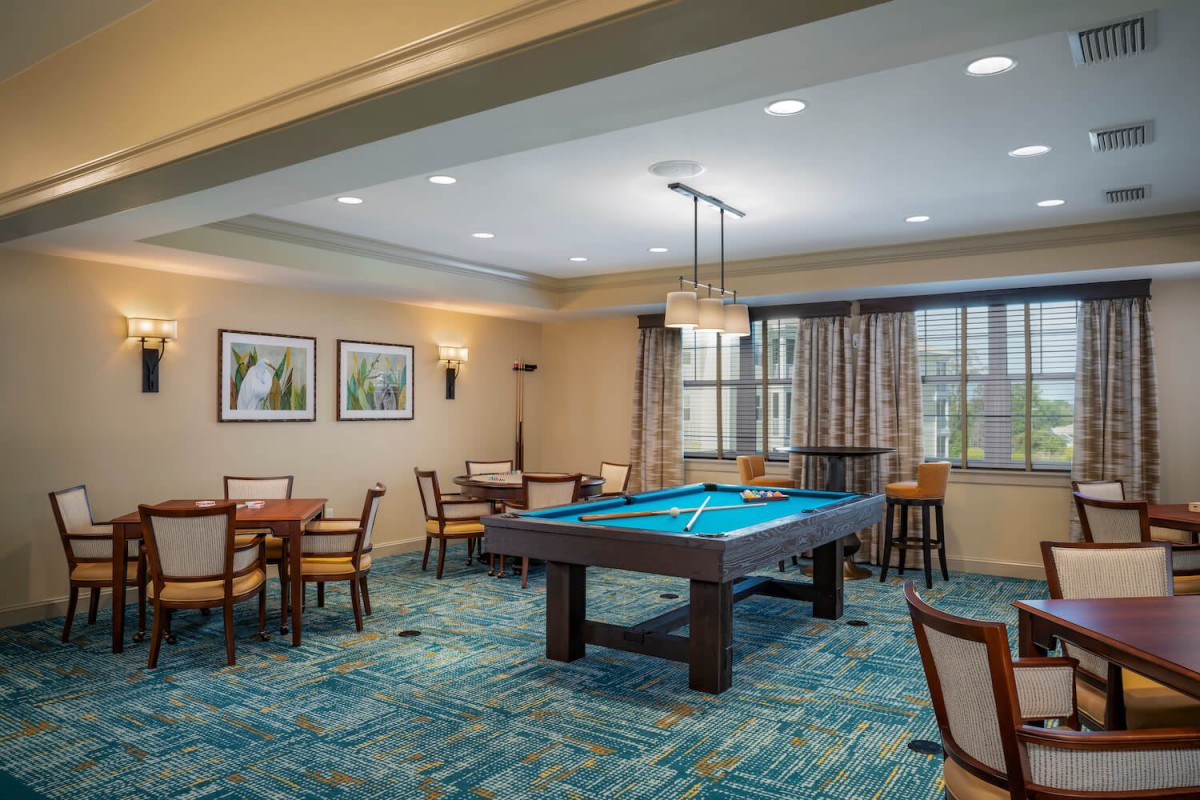 A well-lit game room featuring a billiards table surrounded by several chairs and tables, with decorative elements like framed art and curtains.