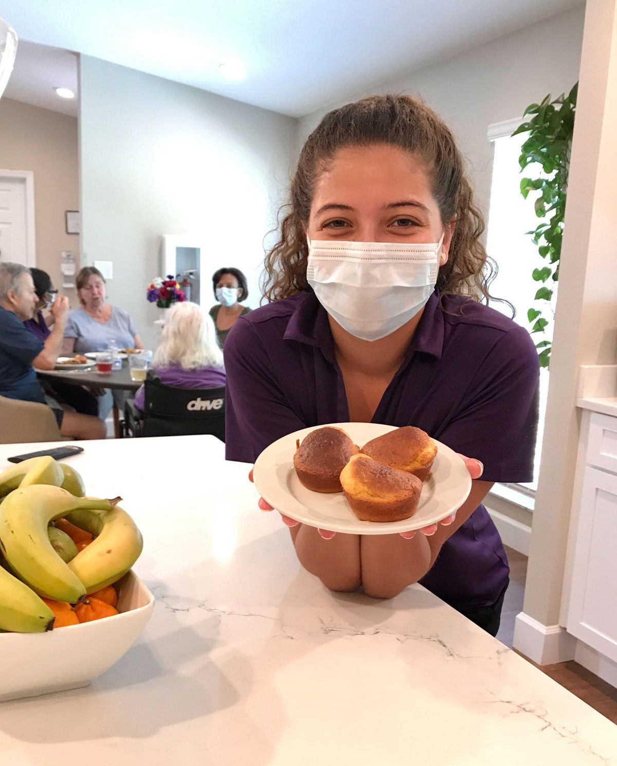 A woman wearing a mask is smiling and holding a plate of buns in a kitchen setting.