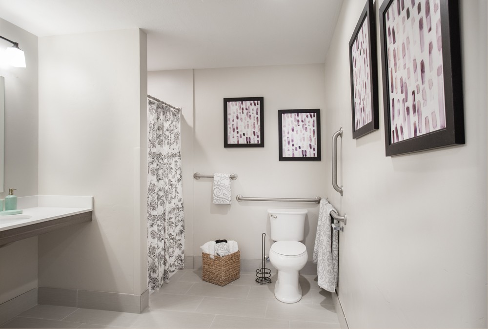 A modern bathroom with neutral tones, featuring a toilet, shower with patterned curtain, decorative wall art, and a towel rack.
