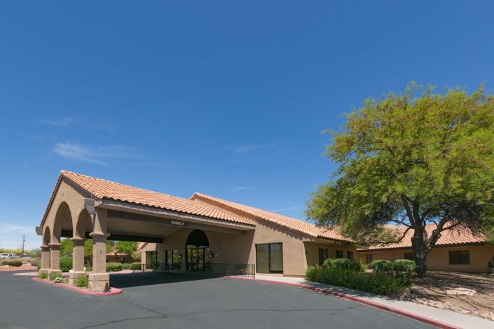 A single-story building with a tiled roof and arched entrance, surrounded by trees and an empty parking lot under a clear blue sky.