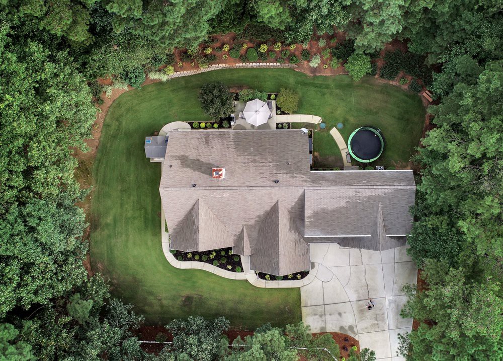 Aerial view of a house surrounded by trees, featuring a well-maintained lawn, a trampoline, a patio area, and a driveway.