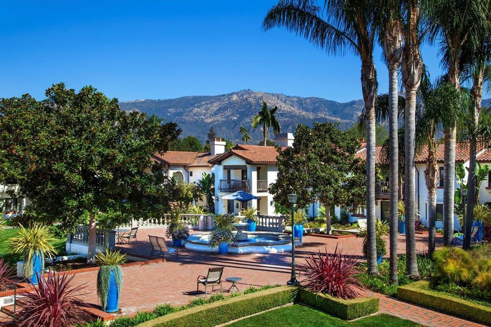 A luxurious resort with Mediterranean-style architecture surrounded by lush landscaping and palm trees, set against a backdrop of mountains.