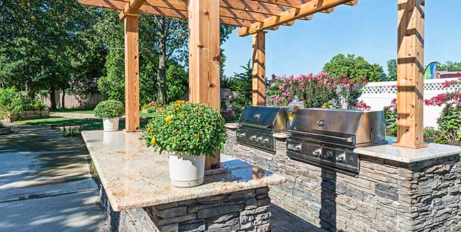 A beautifully designed outdoor kitchen featuring stone countertops and a pergola, complete with multiple grills and surrounded by lush greenery.