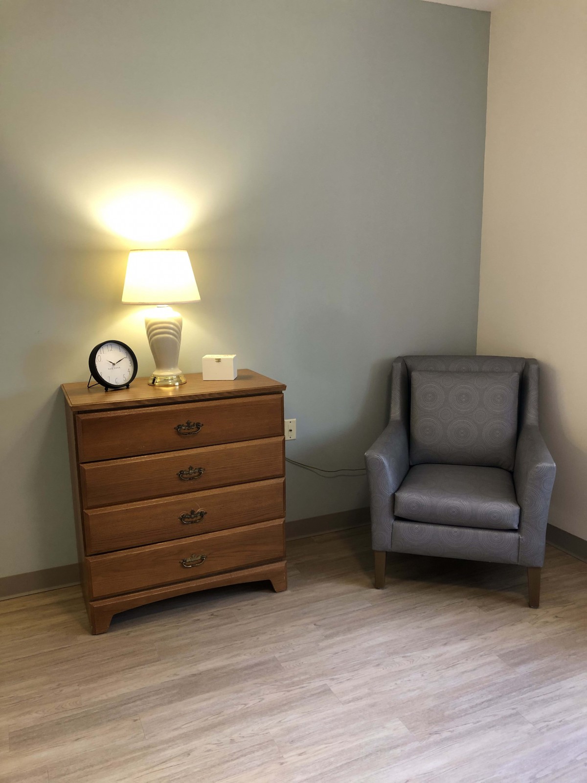 A cozy corner featuring a wooden dresser with a lamp and a comfortable armchair.