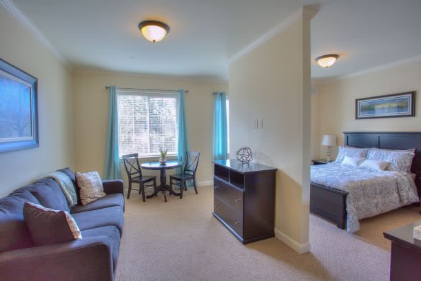 A cozy living space with a dining table, sofa, and an adjacent bedroom, illuminated by natural light through the windows.