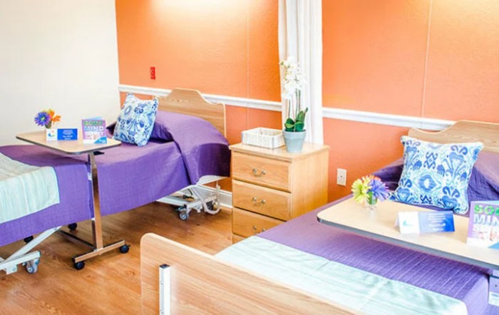A well-lit hospital room with two beds, featuring purple bedding and an orange accent wall.