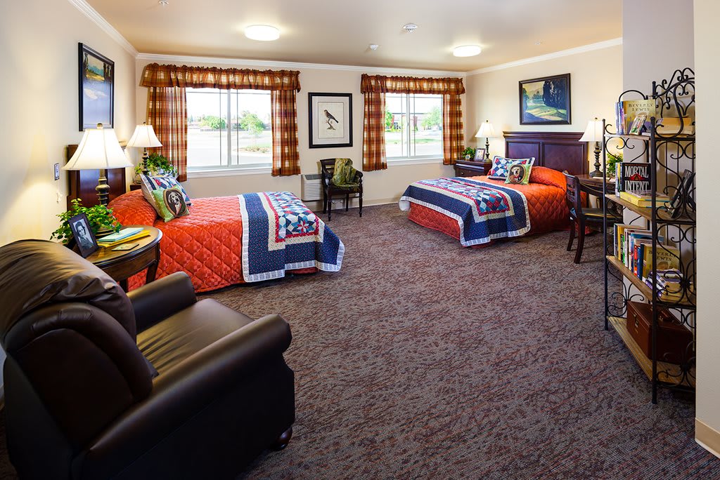 A cozy hotel room featuring two twin beds, colorful quilts, and a carpeted floor, illuminated by natural light from large windows.