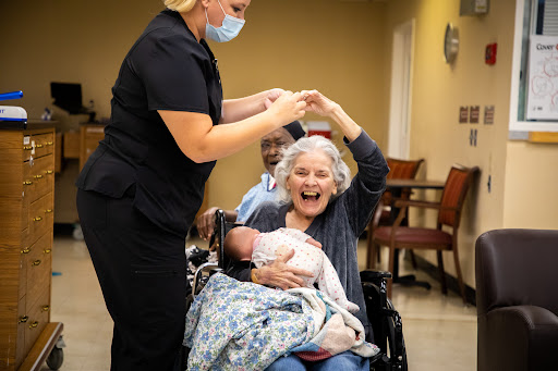 A heartwarming moment as a caregiver celebrates with an elderly woman holding a baby, showcasing the joy of connection and care.