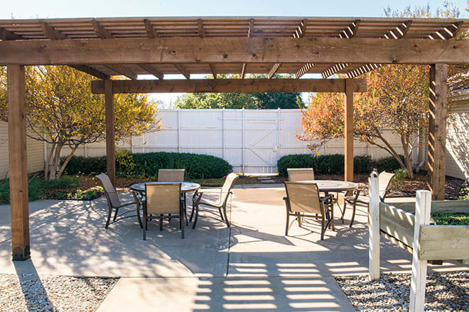 A sunny patio area featuring a wooden pergola, tables, and chairs surrounded by greenery.