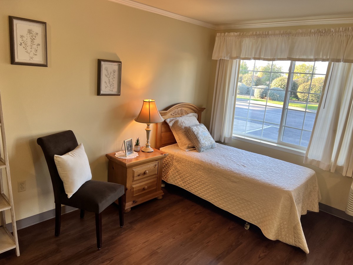 A cozy bedroom with a single bed, a nightstand with a lamp, a chair, and a large window letting in natural light.