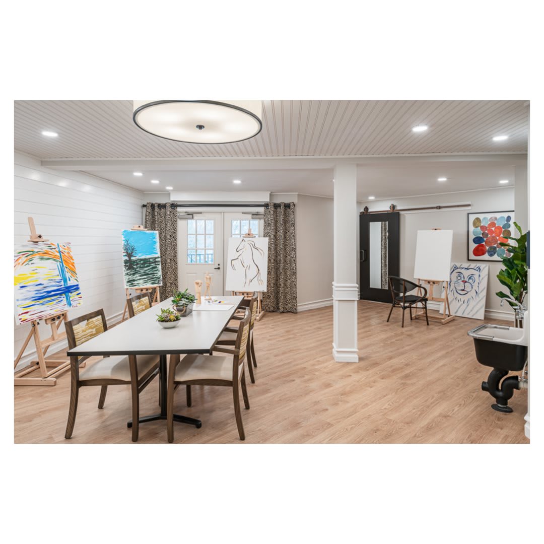 A spacious and modern art studio with a dining area featuring a large table, several chairs, and easels displaying colorful paintings.