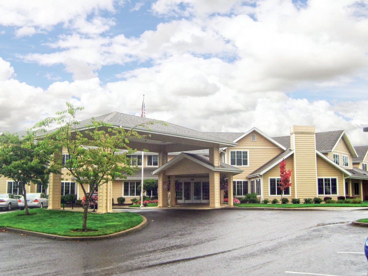 A large, modern building with a covered entrance and surrounding greenery under a partly cloudy sky.