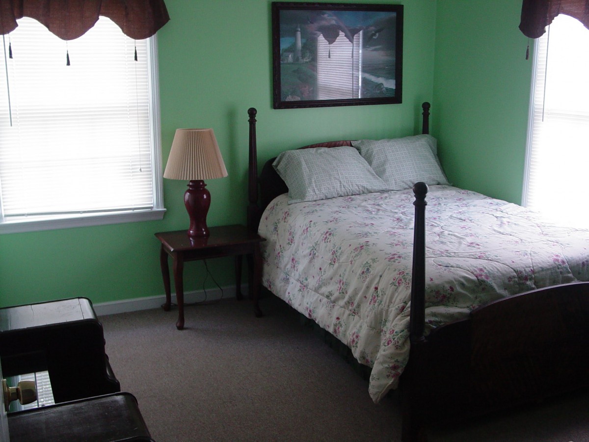 A cozy bedroom with green walls, featuring a bed, nightstand with a lamp, and a window.