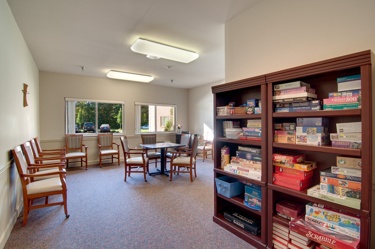A community room with shelves of books and games, surrounded by tables and chairs near large windows.