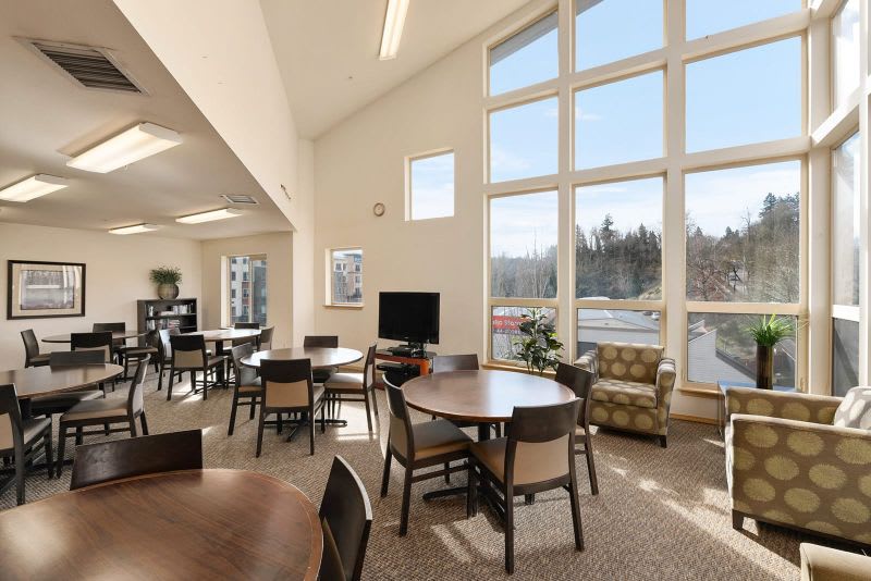 A bright, modern room with large windows, featuring tables and chairs arranged around a television.