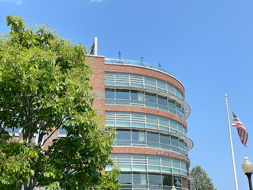 A modern curved building featuring glass windows, surrounded by greenery under a clear blue sky.