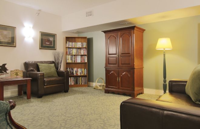 A cozy living room featuring an armchair, wooden cabinet, bookshelf, and a floor lamp on a carpeted floor.