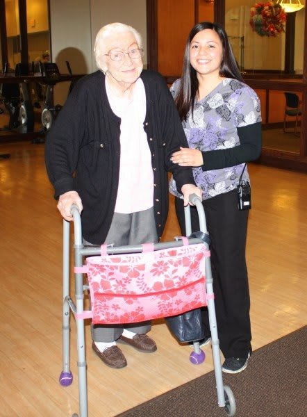 An elderly person with a walker stands next to a caregiver indoors, both smiling.