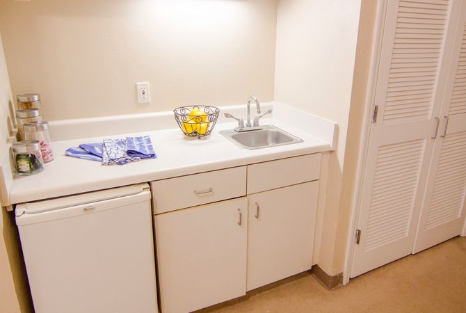 A small kitchenette with a sink, countertop, and fridge, featuring a bowl of lemons and a towel.