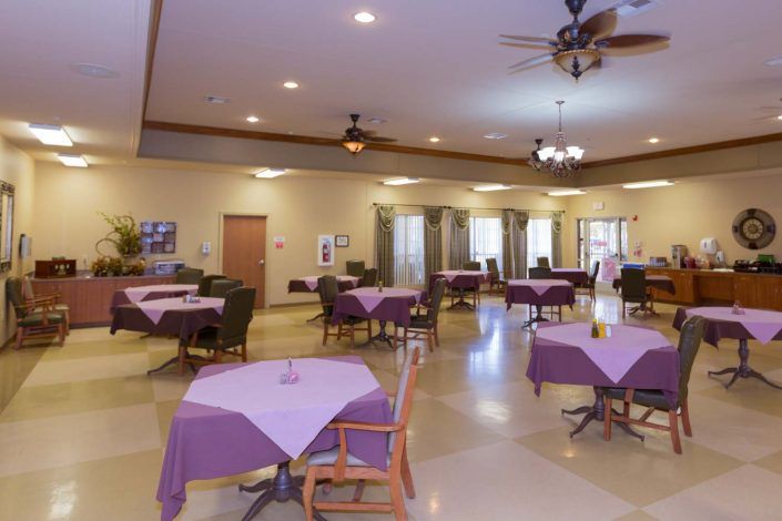 A spacious dining room with tables covered in purple tablecloths, surrounded by chairs and illuminated by ceiling lights and fans.