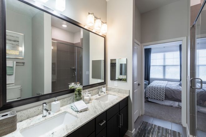 A modern bathroom with a double sink and large mirror, showcasing sleek interior design and a glimpse into an adjoining bedroom.