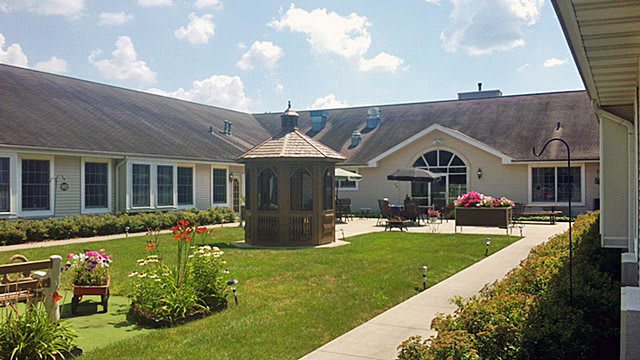 A scenic outdoor area featuring a well-maintained lawn, garden, and gazebo set against a backdrop of residential-style buildings under a partly cloudy sky.