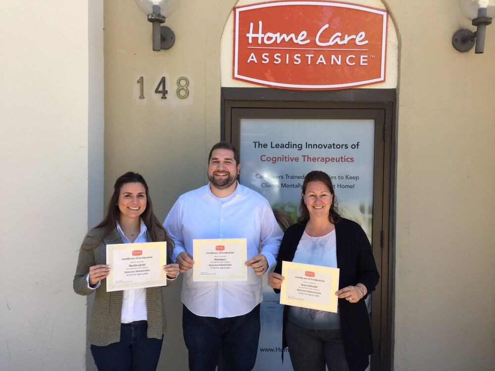 Three individuals proudly hold certificates outside the Home Care Assistance office.