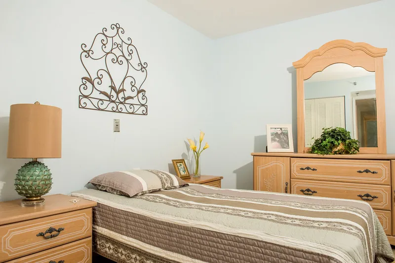A cozy bedroom featuring a neatly made bed, wooden dresser with a mirror, and decorative elements such as flowers and wall art.