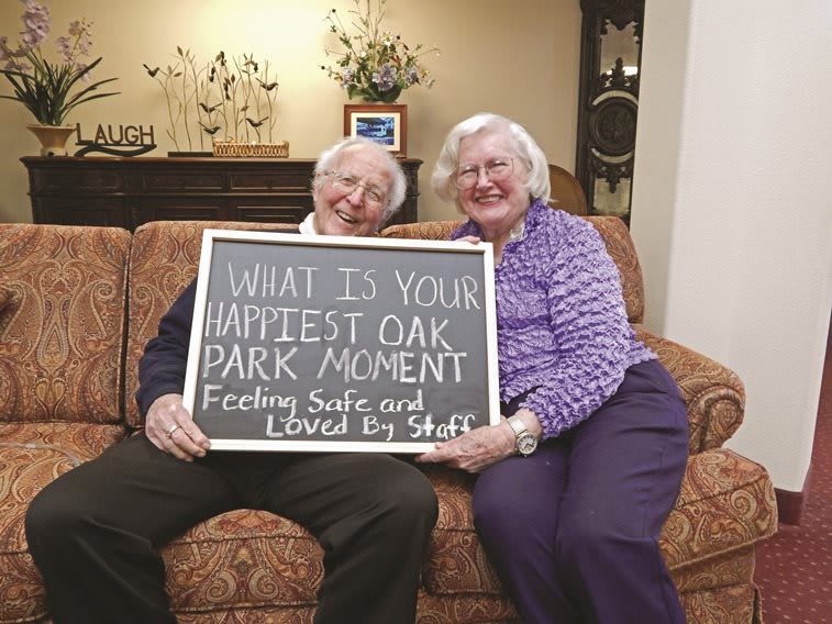 An elderly couple is sitting on a couch, holding a chalkboard sign that reads 'What is your happiest Oak Park moment? Feeling safe and loved by staff.'