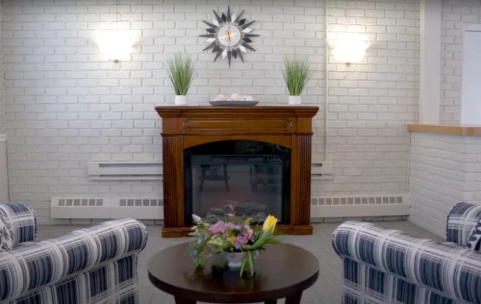 A cozy living room featuring a wooden fireplace with decorative plants, surrounded by striped chairs and a round coffee table with a floral arrangement.