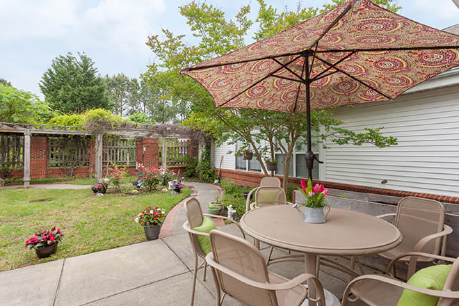 A cozy backyard patio with a table and umbrella, surrounded by greenery and potted plants.