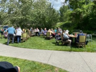 A group of people enjoying an outdoor gathering with tables and chairs arranged on a grassy area under sunny skies.