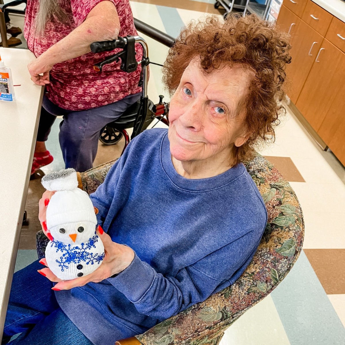 An elderly woman smiling while holding a snowman decoration during an art activity at a community center.