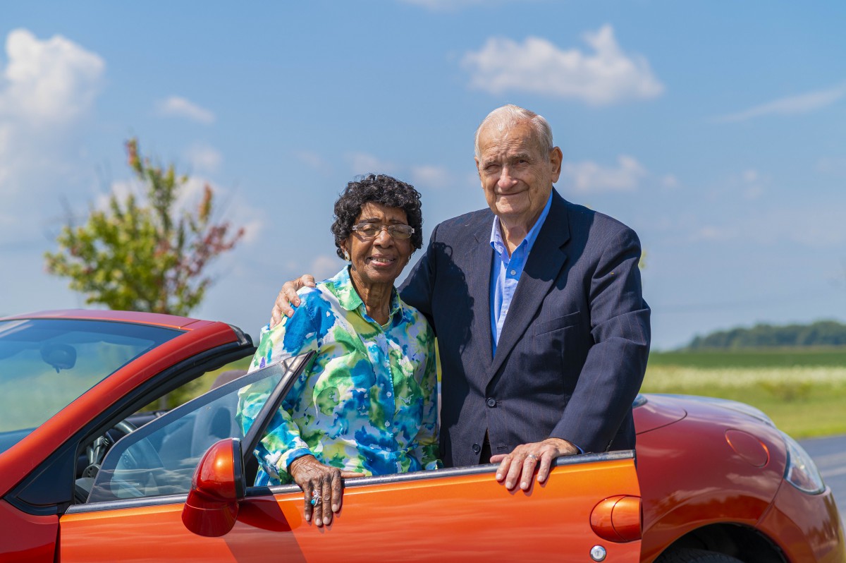 A happy couple poses by their car on a sunny day.