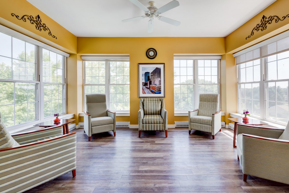 A well-lit room with large windows, featuring several armchairs and wooden flooring.