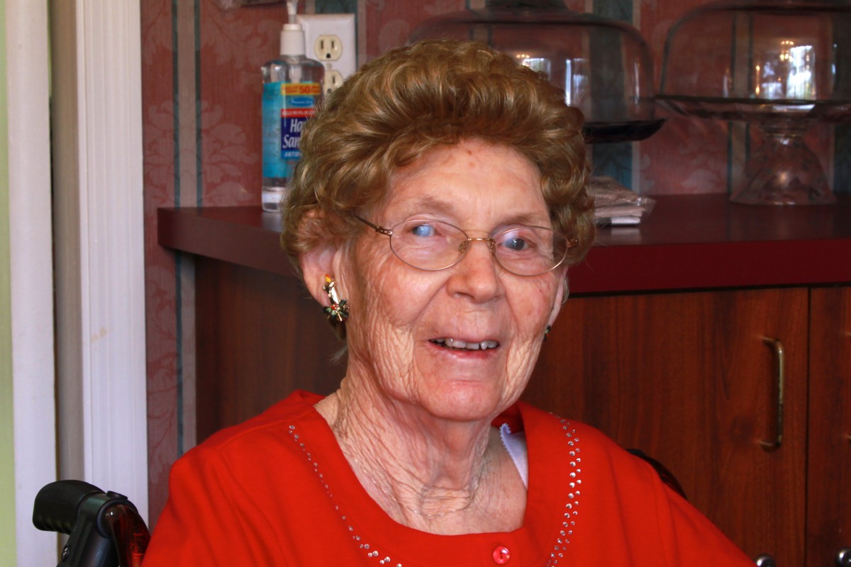 An elderly woman smiling indoors, wearing red clothing.