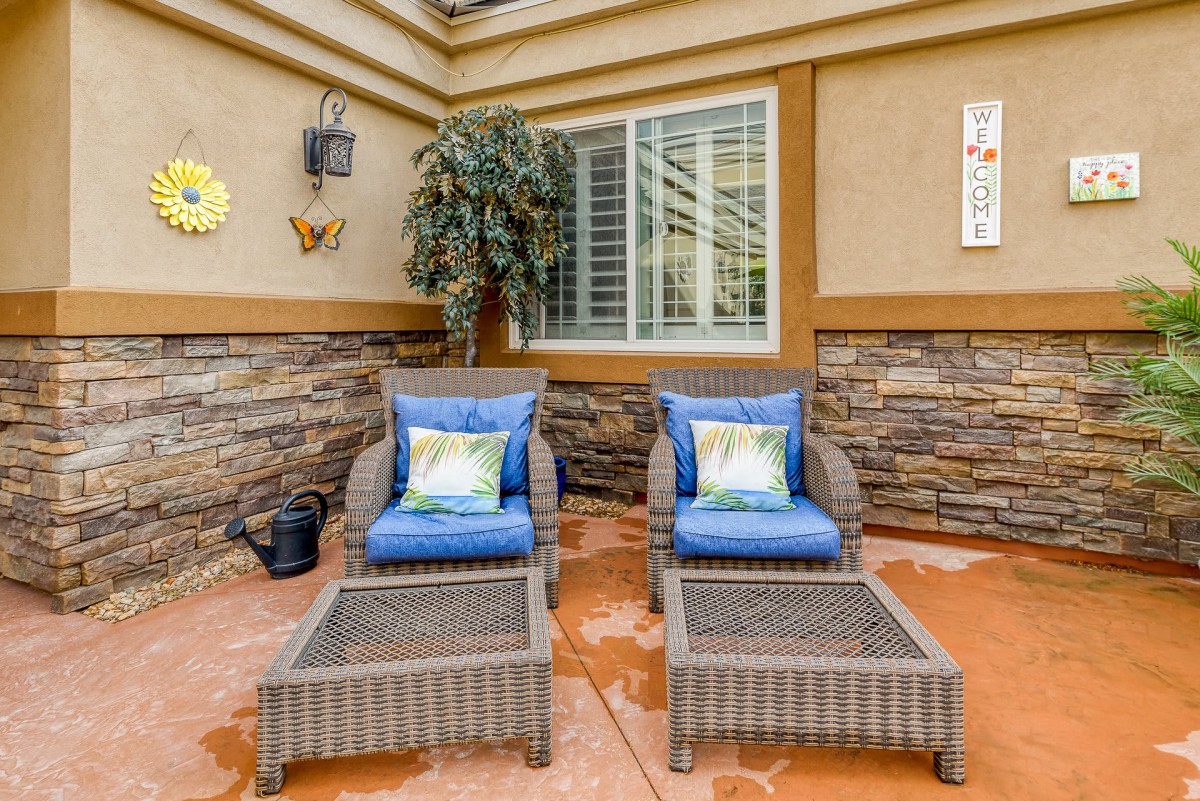 A cozy outdoor patio with wicker chairs featuring blue cushions, positioned against a stone-accented wall and a window, adorned with decorative elements and plants.