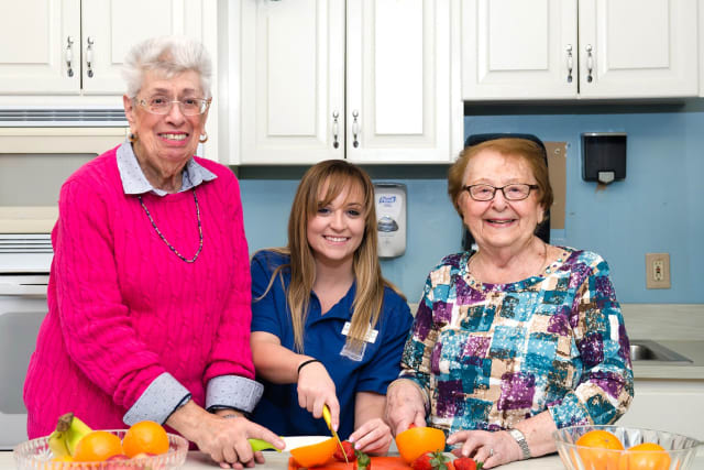 A caregiver and two elderly women are smiling while preparing vegetables in a kitchen.