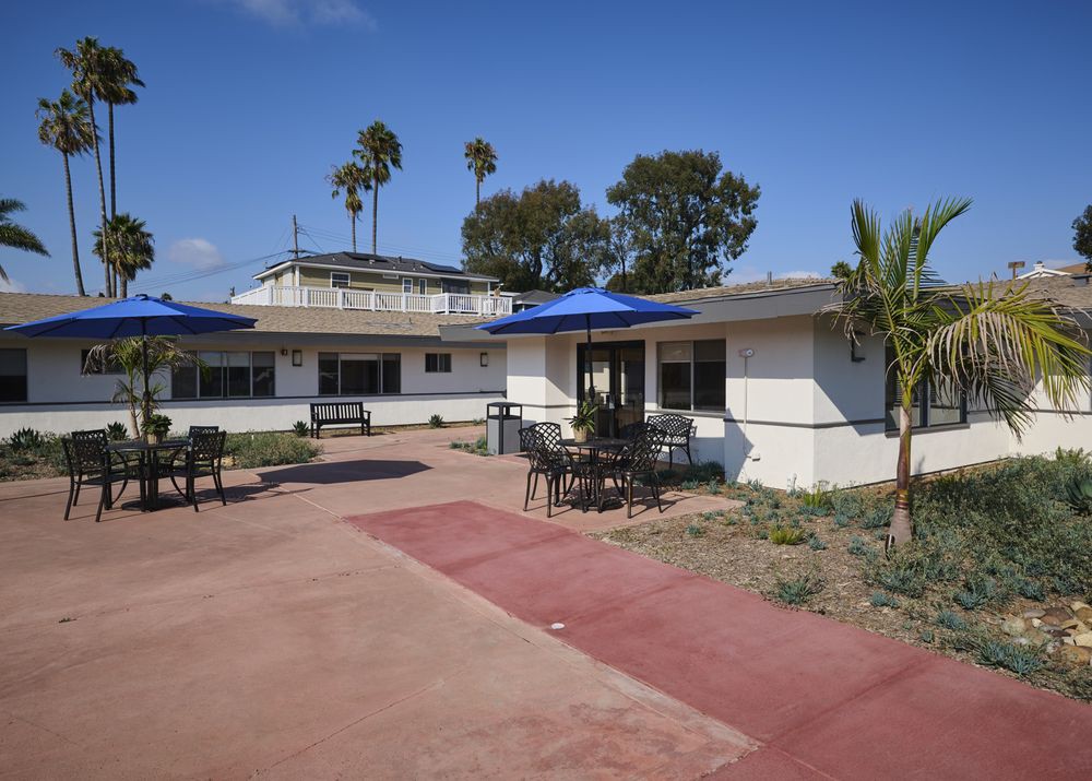 A sunny day at a house with a patio and blue umbrellas, surrounded by palm trees.