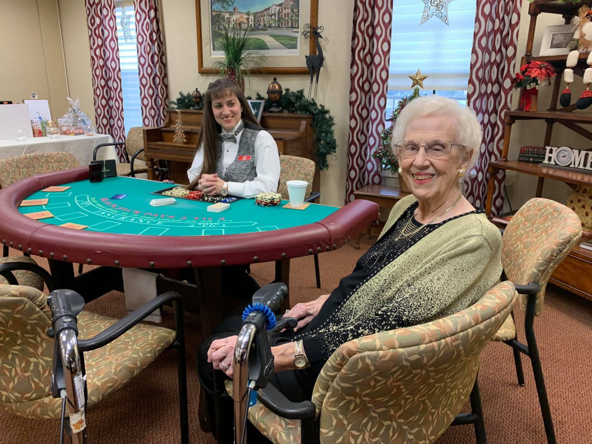 An elderly woman smiling while seated at a card table, ready to play.