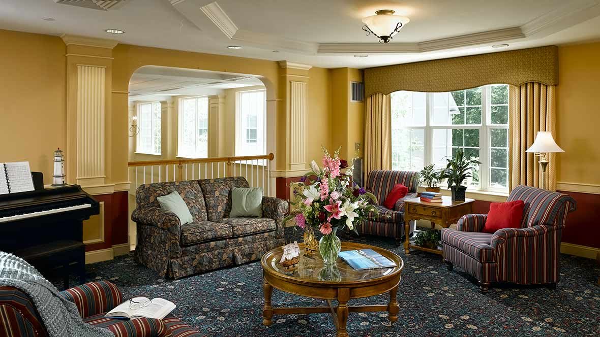 A cozy living room featuring patterned sofas, a floral arrangement on a round coffee table, and large windows with curtains.
