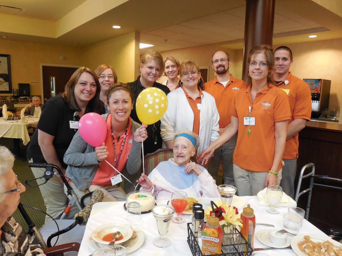 A cheerful group of people celebrating with an elderly person at a dining table, decorated with colorful balloons.