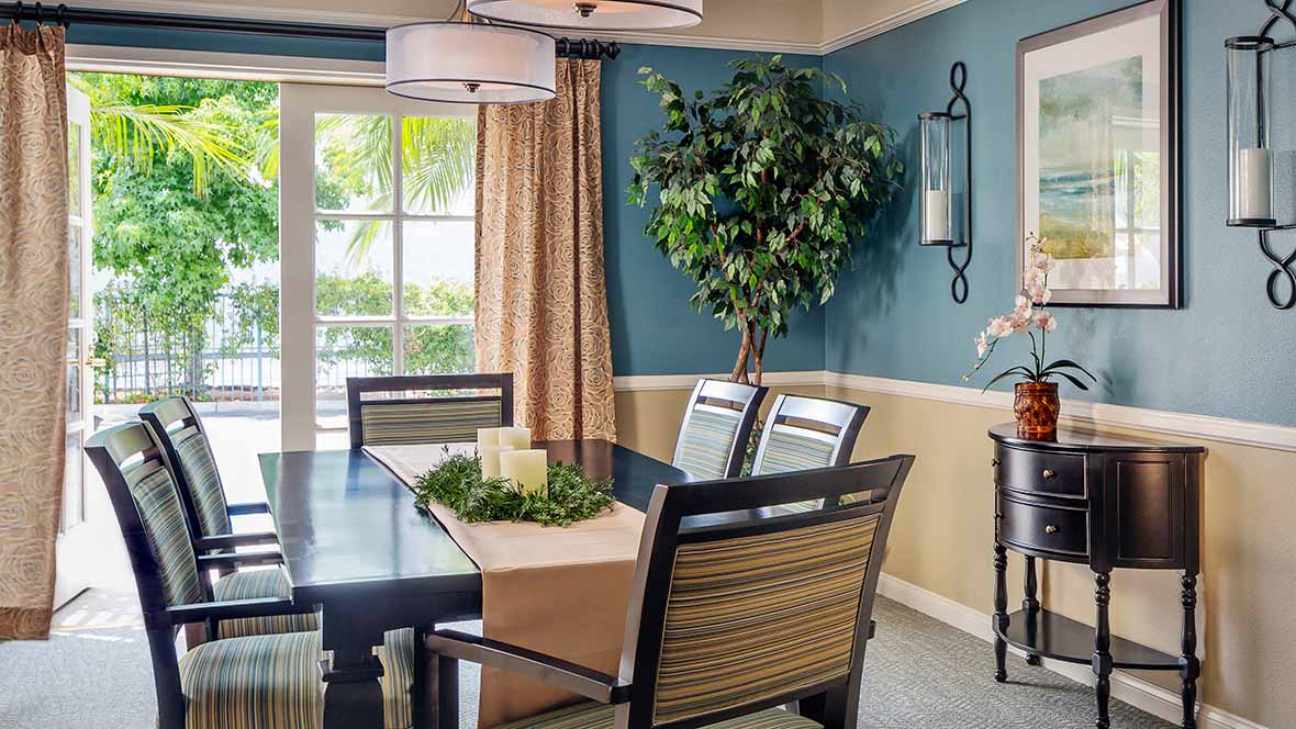 A cozy dining room with a table set for four, featuring a green plant and natural light from a large window.