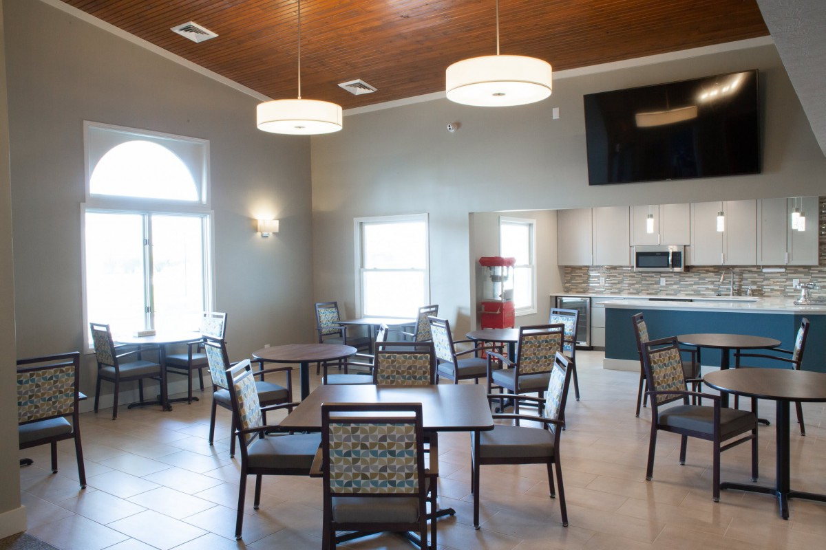 A modern community dining area with several tables and chairs, featuring a kitchen in the background.