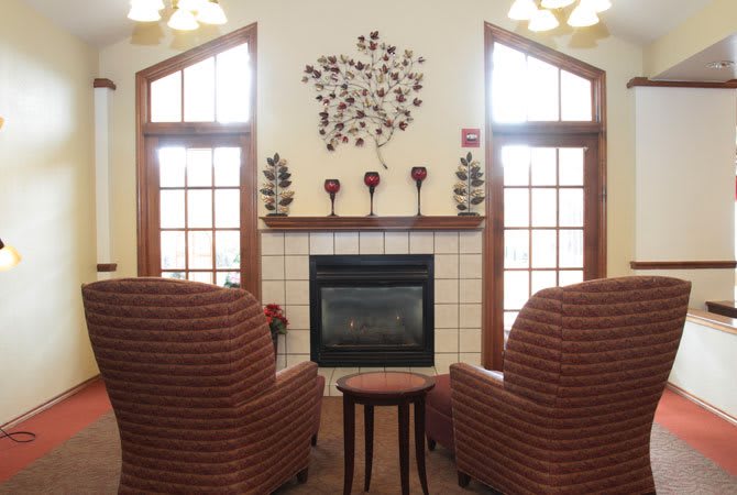 A cozy living room with two armchairs facing a fireplace, adorned with decorative wall art and flanked by large windows.